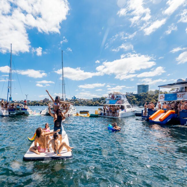 A lively scene of people enjoying a sunny day on the water with various boats anchored nearby. Some individuals are on floating platforms, others are swimming, and a few are on inflatables. The backdrop includes clear blue skies and distant buildings, courtesy of The Yacht Social Club Sydney Boat Hire.