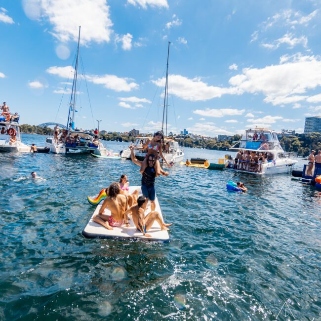 A lively scene on a sunny day with people enjoying the water on paddleboards and around yachts. Some are swimming, while others are relaxing and socializing on anchored boats. The background features a clear blue sky and scattered clouds, perfect for The Yacht Social Club Event Boat Charters.