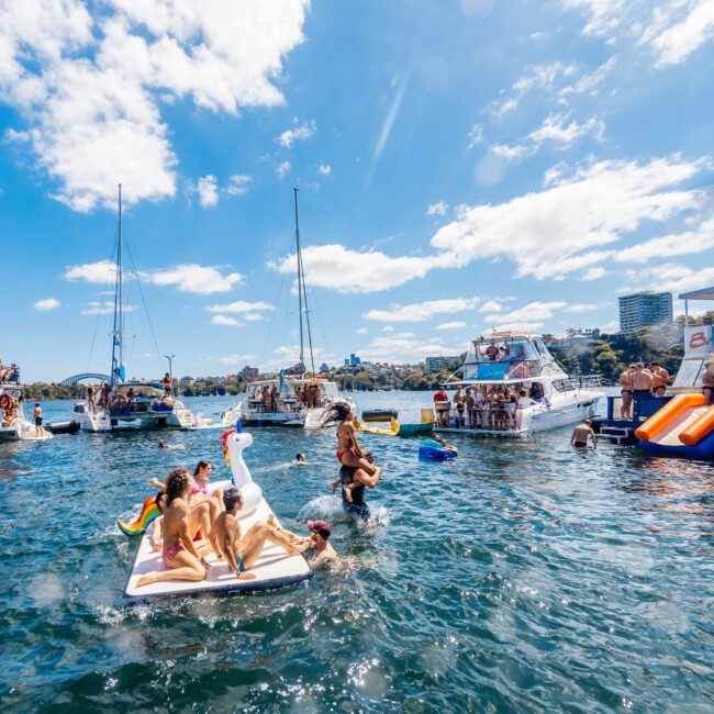 People are enjoying a sunny day on a lake surrounded by boats and inflatables. Some individuals lounge on a large inflatable swan, while others swim in the water. The scene is lively, with people socializing and engaging in activities reminiscent of The Yacht Social Club's boat parties in Sydney Harbour.