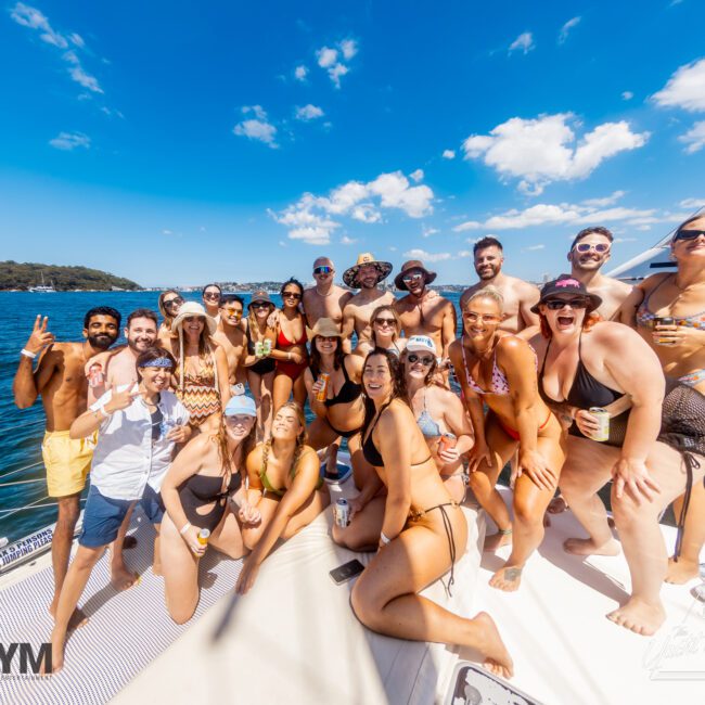 A large group of people gathered on a boat deck, smiling and posing for the camera under a clear blue sky. They are dressed in swimsuits, enjoying a sunny day on the water. In the background, lush green trees and Sydney Harbour enhance the picturesque scenery at a Yacht Social Club event.