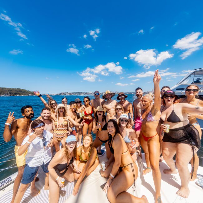 A group of people celebrating on a boat under a clear blue sky. Most are wearing swimwear and smiling at the camera, some with drinks in hand. The boat is on a body of water with visible shoreline in the background. Logos "GAYM" and "Jackson Grant Photography" are visible. The Yacht Social Club Sydney Boat Hire is mentioned prominently.