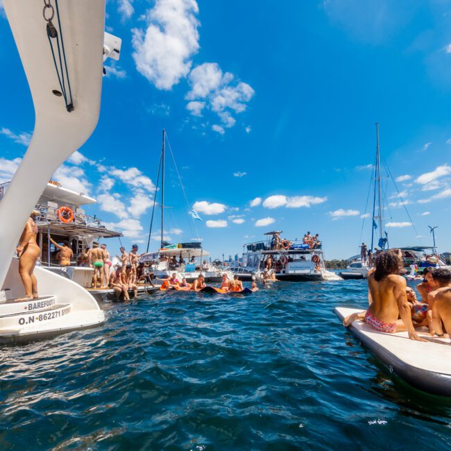 People are enjoying a sunny day on the water, swimming and lounging on paddleboards among several anchored boats. The clear sky and deep blue water create a vibrant, festive atmosphere, perfect for luxury yacht rentals from The Yacht Social Club in Sydney Harbour.