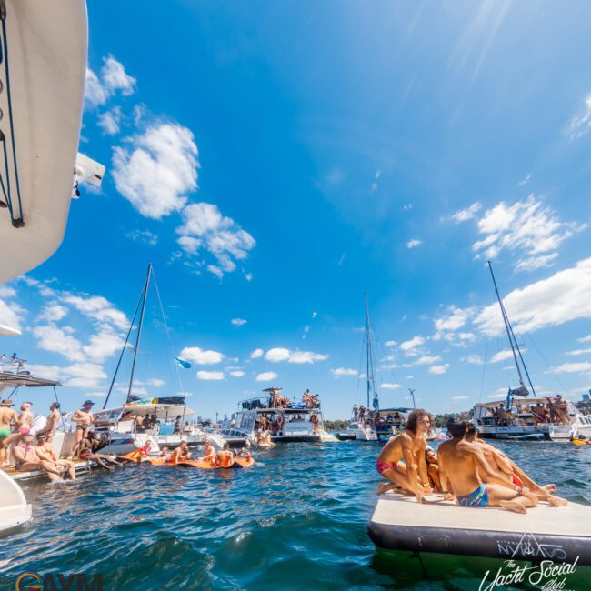 This image shows a lively scene on the water with several boats docked close together under a bright blue sky with scattered clouds. People are enjoying themselves, swimming, and lounging on floating platforms. Logos for The Yacht Social Club and Boat Parties Sydney are also visible.