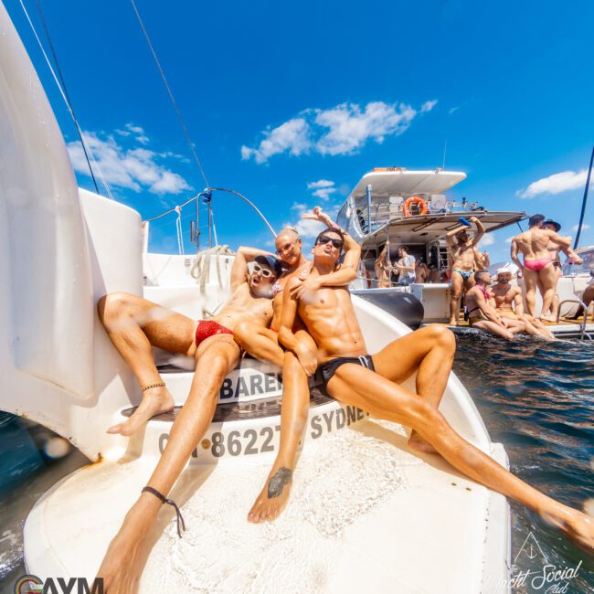 Three people in swimwear are relaxing on the back of a luxury yacht, enjoying a sunny day on the water. They are posing cheerfully with other people and boats visible in the background. The sky is clear and blue, perfect for Boat Parties Sydney The Yacht Social Club.