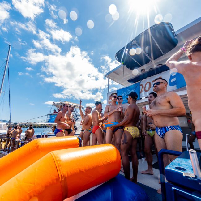 A lively group of people in swimsuits gather on a boat under a sunny sky. There are inflatable orange water slides and other boats nearby. The festive mood is enhanced by bright colors and clear water. Logos for "GAYM" and "Luxury Yacht Rentals Sydney" are visible in the corners.