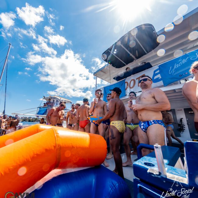 A group of people in swimsuits stand on a boat's deck under a sunny sky. Some hold drinks, with inflatable pool toys visible in the foreground. A banner with "The Yacht Social Club" is displayed above them, highlighting the fun at Sydney Harbour Boat Hire The Yacht Social Club.