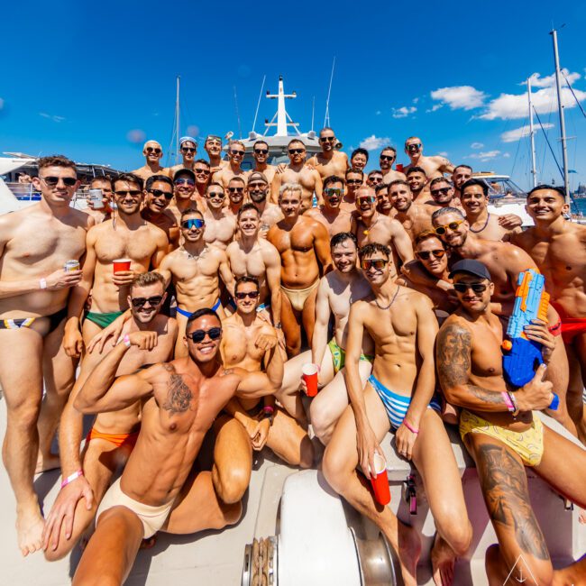 A large group of men wearing swimwear and sunglasses poses together on a yacht under a bright blue sky. Some are holding drinks, and one person holds a water gun. The festive scene is lively, with yachts visible in the background. Logos in the corners read "GAY M" and "The Yacht Social Club Sydney Harbour Boat Hire.