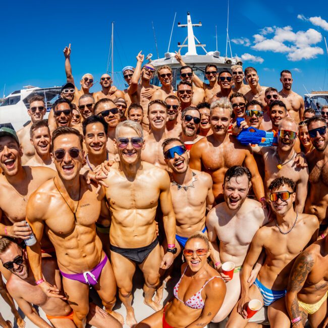 A large group of men are gathered on a boat, most of them shirtless and wearing swimwear. They are smiling and posing for the camera, with clear blue skies and other boats in the background. Some hold drinks and sunglasses, indicating a festive atmosphere at The Yacht Social Club Event Boat Charters.