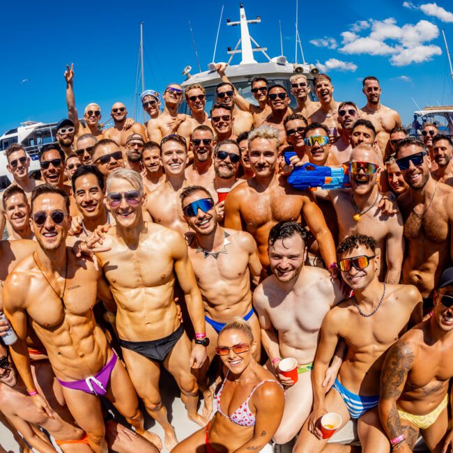 A large group of smiling people, mostly men, gather shirtless in colorful swimwear on a boat under a sunny sky. Some wear sunglasses, hold drinks, and appear to be enjoying a lively atmosphere at The Yacht Social Club. Several yachts and the city skyline are visible in the background of Sydney Harbour.