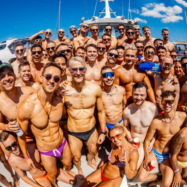 A large group of people, mostly shirtless men in swimwear, is gathered on the deck of a boat under a clear, sunny sky. Many are smiling and wearing sunglasses. In the background, city skyscrapers and another boat can be seen on the water—a perfect day for Boat Parties Sydney The Yacht Social Club.