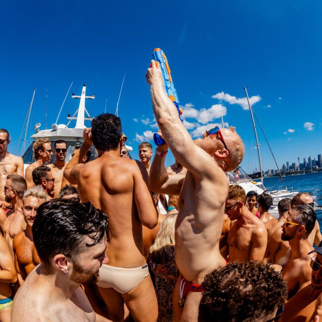 A lively group of men in swimwear enjoying a boat party on a sunny day. One man stands out, holding a colorful water gun, spraying water upwards. The backdrop features a city skyline with skyscrapers and a clear blue sky, courtesy of Boat Parties Sydney The Yacht Social Club.