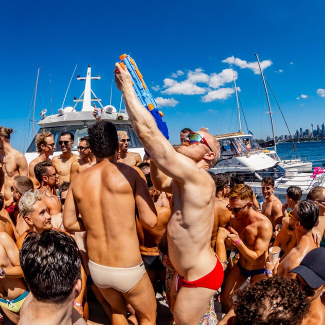 A lively group of people in swimwear are celebrating on a boat with a city skyline in the background. One person in red trunks is holding a large water gun, with others around them cheering and having fun under a clear, sunny sky at The Yacht Social Club Event by Sydney Harbour Boat Hire.