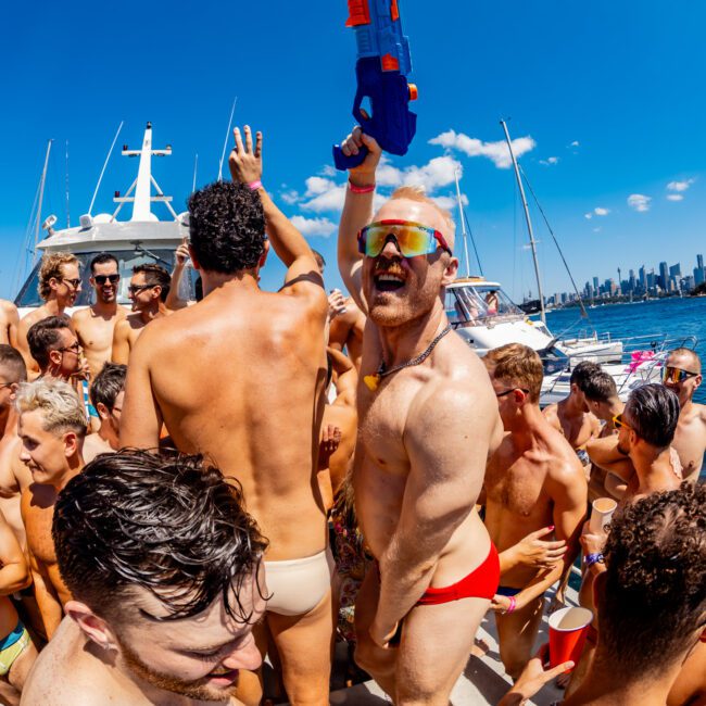 A lively party scene on a yacht with numerous shirtless individuals in swimwear. One person in red swim trunks and sunglasses joyfully holds up a blue water gun. The sky is clear, and the city skyline is visible in the background, capturing the essence of Boat Parties Sydney The Yacht Social Club.