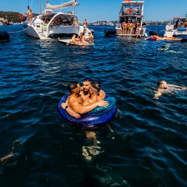 People are enjoying a sunny day on the water, some floating on inflatable devices and others swimming. Boats are anchored in the background, and several individuals are relaxing on them. In the foreground, a group of three people is sitting on a circular blue float at The Yacht Social Club Event.