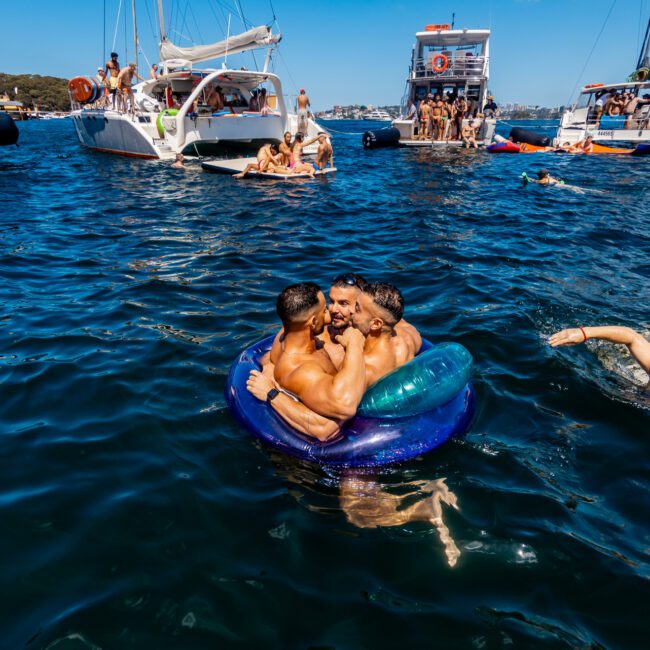 Three men are sitting closely together in a small inflatable ring on the water, embracing and laughing. They are surrounded by several yachts and people swimming or lounging on the boats from The Yacht Social Club Sydney Boat Hire. The event appears festive and social under a clear blue sky.