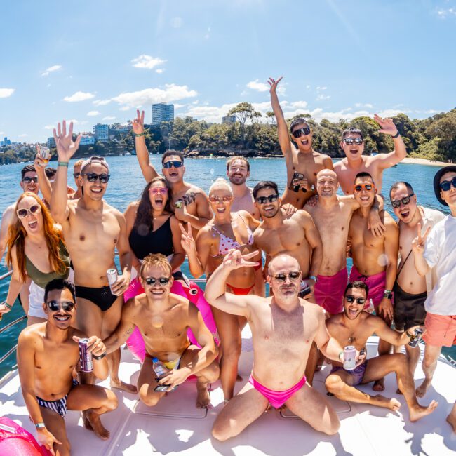 A cheerful group of around 20 people poses for a photo on a boat. They are wearing swimsuits and sunglasses, with many holding drinks and smiling at the camera. The boat, part of The Yacht Social Club Event Boat Charters, is on a body of water with a city skyline and trees in the background under a bright blue sky.