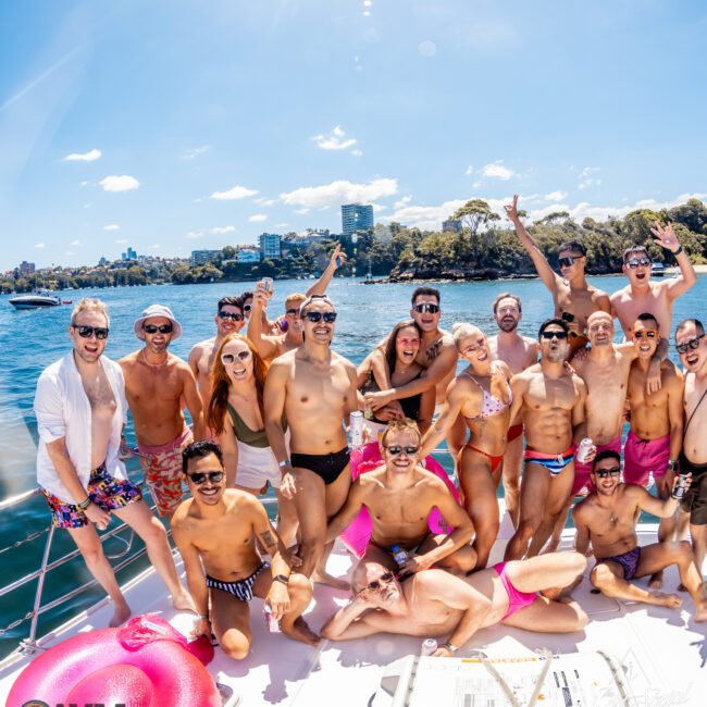 A joyful group of people in swimwear pose for a photo on a sunny day aboard The Yacht Social Club Event. The clear blue sky and water create a vibrant backdrop. Most are smiling or cheering, enjoying the festive atmosphere. An inflatable pink ring is also in the frame.