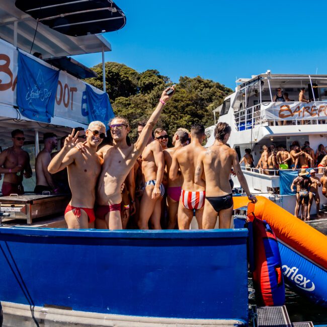 A group of people in swimwear are partying on boats. The boats, provided by The Yacht Social Club, are decorated with banners and inflatable toys, with attendees enjoying the lively atmosphere under the sun. Some individuals are taking selfies, and the water is filled with other partygoers.