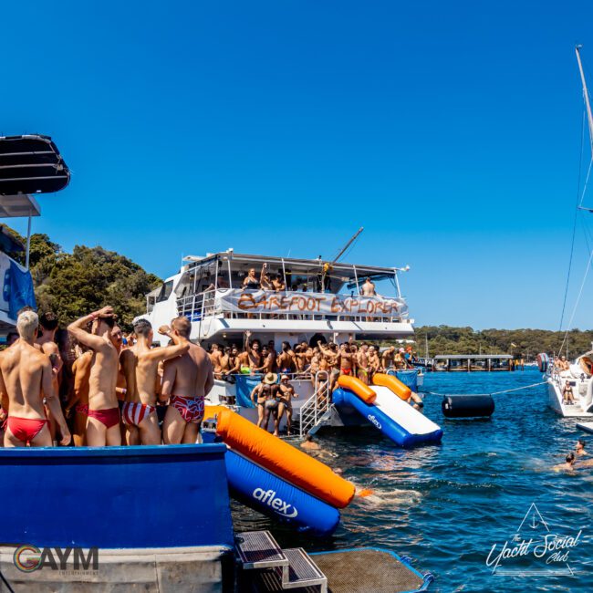 A lively boat party shows a large group of people in swimwear enjoying the sun on a yacht named "Barefoot Explorer." The scene includes inflatable slides, another boat nearby, and clear blue waters. The "GAYM" and "The Yacht Social Club Sydney Boat Hire" logos are visible in the foreground.