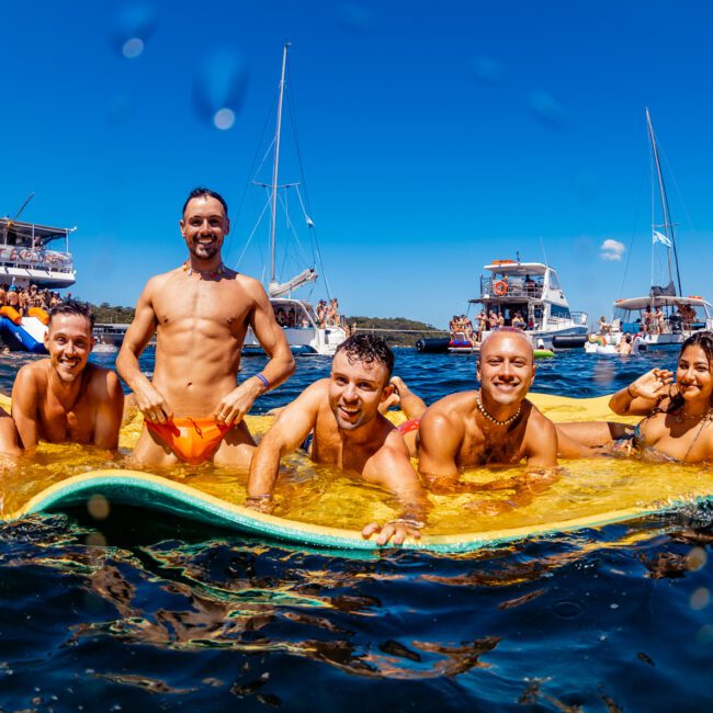 A group of seven people smiling and lying on a large floating mat in the water, with several boats and other people in the background. The scene is vibrant and energetic, suggesting a fun, social event hosted by The Yacht Social Club Sydney Boat Hire. The "Yacht Social Club" logo is visible in the corner.