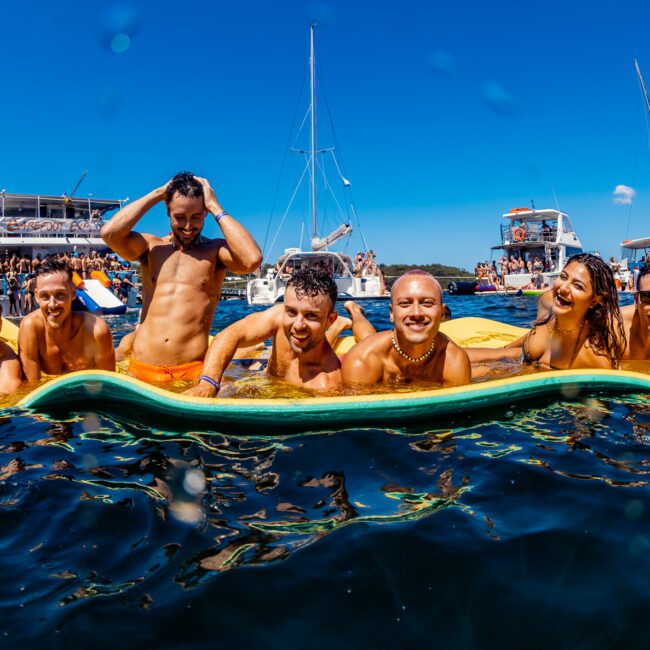 A group of seven smiling people, wearing swimwear, are enjoying themselves on a floating mat in the water. Behind them, several yachts from The Yacht Social Club are anchored, and other groups are socializing under the clear blue sky. The atmosphere is festive and lively.