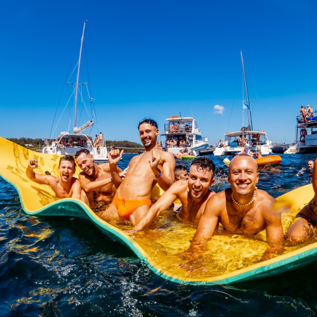 A group of seven people are lying on a yellow floating mat in the ocean, surrounded by several boats. They are smiling and enjoying the sunny day at The Yacht Social Club. Some boats in the background have people on board, and the sky is clear with a few scattered clouds.