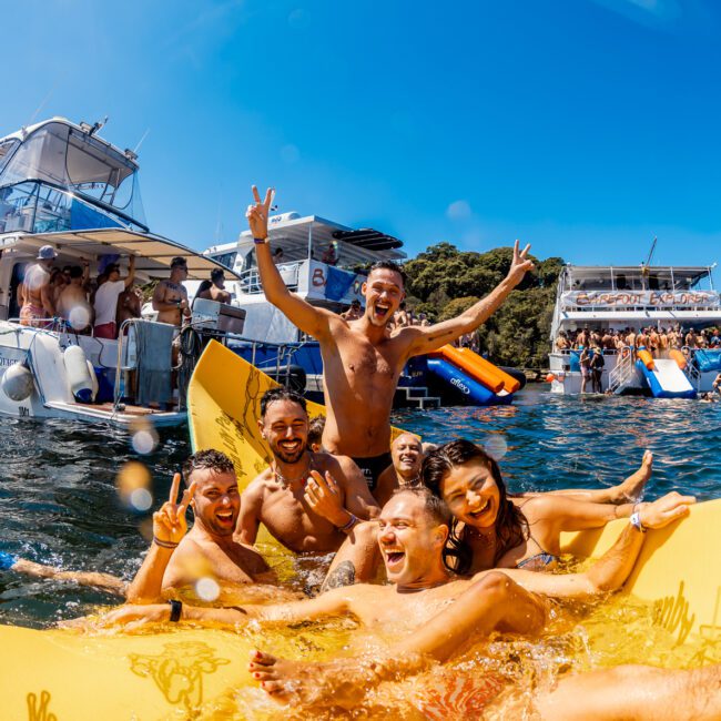 A group of people enjoy a sunny day on the water, smiling and posing on an inflatable raft. Behind them, multiple boats anchor close together, loaded with more people socializing and partying. The water is clear and blue, with a backdrop of lush green trees—an idyllic scene at The Yacht Social Club hosted by Sydney Harbour Boat Hire.