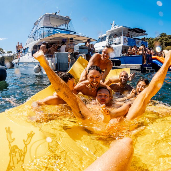 A group of people are lounging and playing on an inflatable yellow water mat in a sunny, outdoor setting. Boats from the Sydney Harbour Boat Hire The Yacht Social Club are anchored around them, with more people socializing on board. The background includes trees and buildings, making for a lively and fun atmosphere.