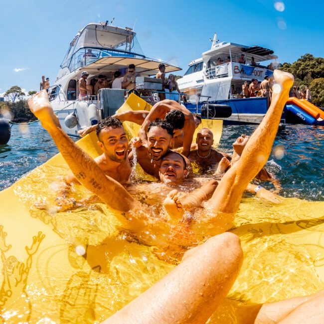 A group of joyful men are lounging on a large, yellow, floating mat in the water during The Yacht Social Club Event Boat Charters. They're smiling, laughing, and raising their arms in celebration under a cloudless, sunny sky with several boats and other people in the background.