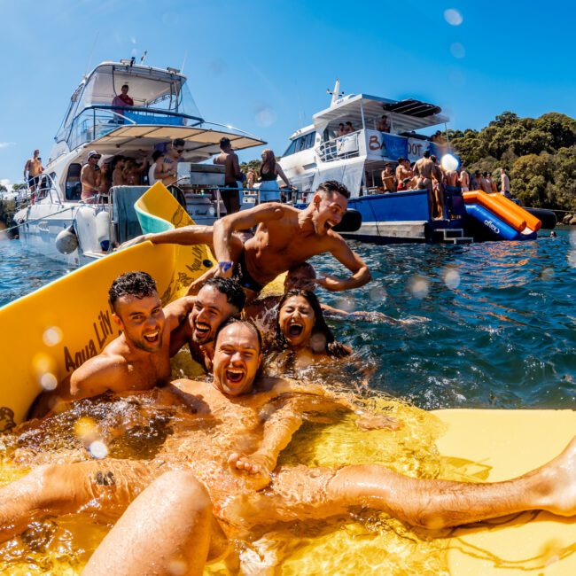 A group of people are having fun on a yellow floating mat in a body of water, near several boats. They are smiling and appear to be enjoying the sun. The background shows other people on boats and water slides. It's a lively scene, perfect for a day with The Yacht Social Club Sydney Boat Hire.