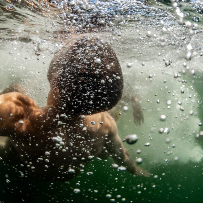 Underwater view of a person in mid-air after jumping into water, with their back visible and bubbles surrounding them. The water appears green, and the person is partially submerged. The image captures the motion and energy of the jump, reminiscent of the fun at Boat Parties Sydney The Yacht Social Club.