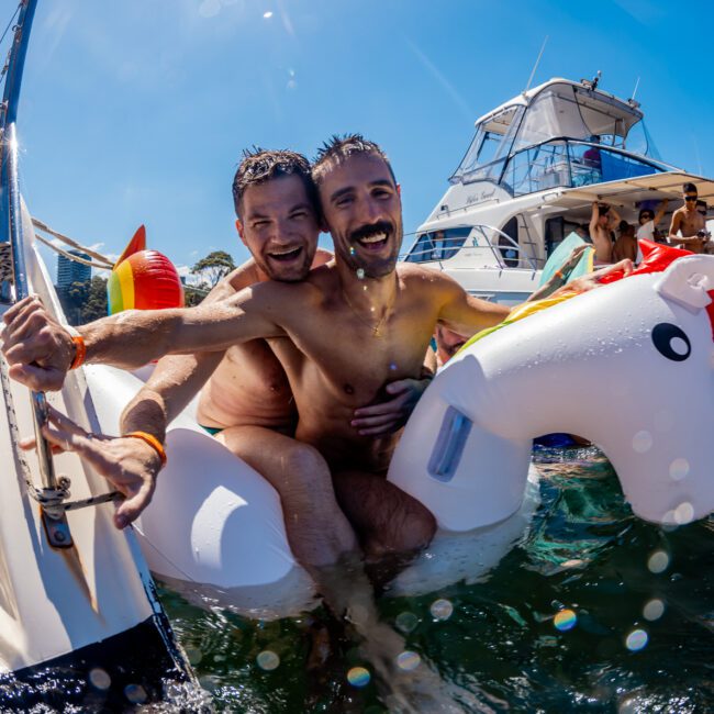 Two men are joyfully riding a white unicorn float near a boat during The Yacht Social Club Sydney Boat Hire event, under a sunny sky. They are surrounded by others enjoying water activities. The atmosphere is lively and festive, with boats and inflatables in the background.