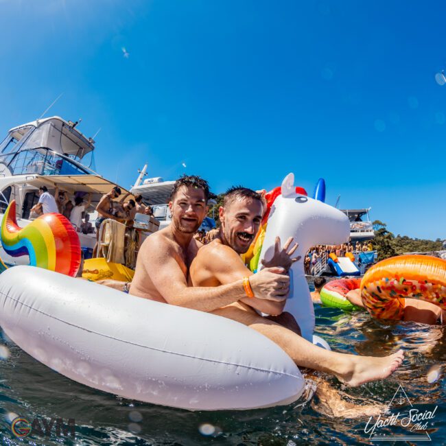 Two happy men sit on an inflatable unicorn pool float in the water, surrounded by various colorful inflatables, including a rainbow and a donut. A yacht is in the background under a clear blue sky, part of The Yacht Social Club’s luxurious boat parties in Sydney, where others enjoy the lively gathering.