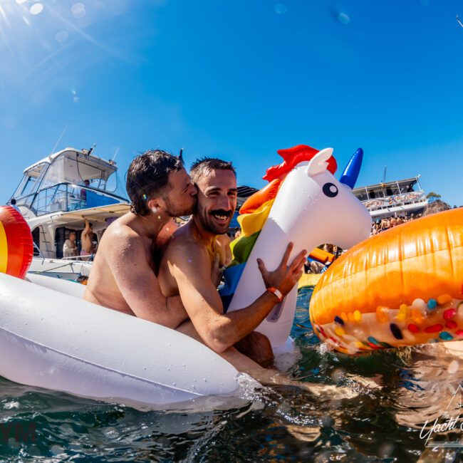 Two men joyfully embrace on inflatable pool toys in the water, with one kissing the other on the cheek. They're surrounded by boats and people under a bright blue sky. One inflatable is a unicorn, and another is a donut. A watermark reads "Boat Parties Sydney The Yacht Social Club.”