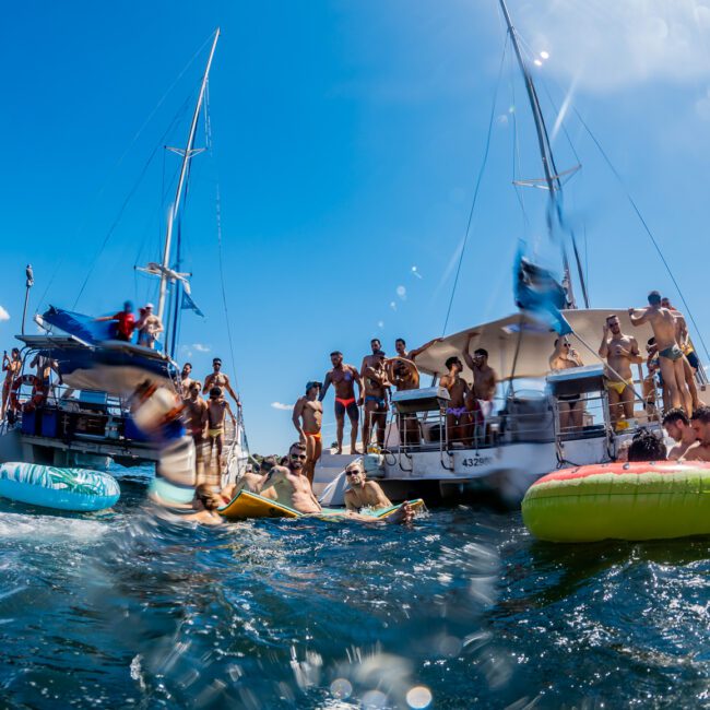 People are enjoying a sunny day on the water, swimming and floating on inflatables near several boats. Some are on the boats while others are in the water, creating a lively and festive atmosphere under a clear blue sky. The scene is framed by the coast in the background, featuring Luxury Yacht Rentals Sydney.