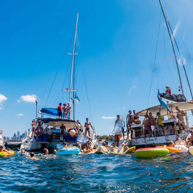 Boats are anchored in a sunny, crowded bay, with many people swimming and floating on inflatables. Some participants enjoy The Yacht Social Club Event Boat Charters, socializing and taking in the weather. The blue sky is clear, and the water looks inviting. The scene is lively and festive.