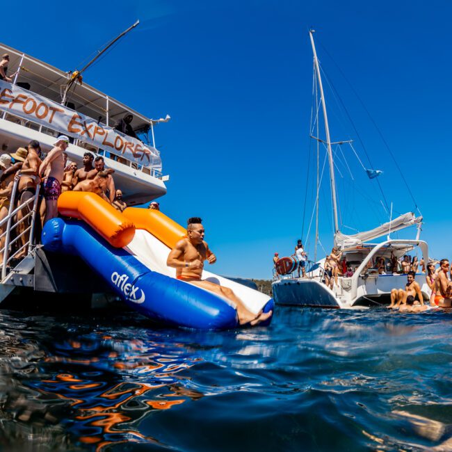 Numerous people are enjoying a sunny day on and around a large boat with a slide into the water. Some are on the boat's deck, while others swim or relax on smaller boats nearby. The boat has a sign reading “Barefoot Explorers.” This vibrant scene embodies The Yacht Social Club Sydney Boat Hire experience under clear blue skies.