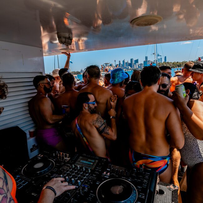 A group of people are gathered on a boat, enjoying a lively party hosted by Boat Parties Sydney The Yacht Social Club. Many are wearing swimwear and sunglasses, some holding drinks. A DJ is performing on the left side with decks and equipment. In the background, more boats and a city skyline under a clear blue sky complete the scene.