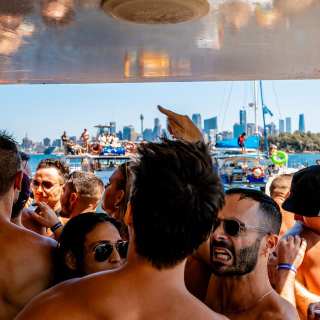 A crowd of people enjoying an outdoor boat party under a clear sky, with many shirtless individuals facing various directions. In the background, more boats and the skyline of a city, including tall buildings and a bridge, can be seen. Experience this vibrant scene with The Yacht Social Club Sydney Boat Hire.