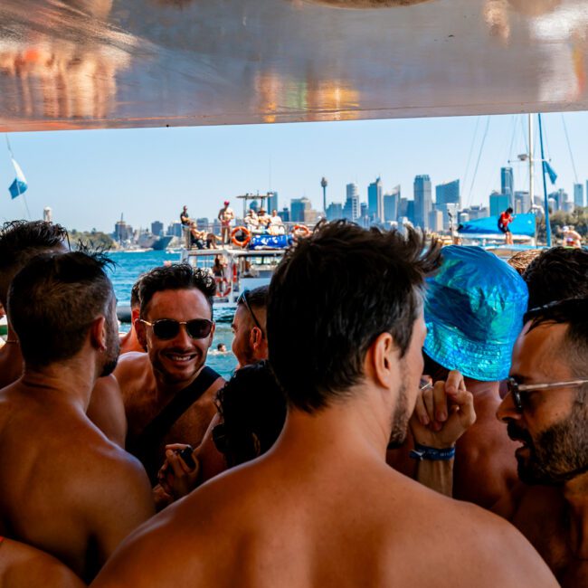 A large group of shirtless men are gathered closely together on a boat under a canopy on a sunny day. In the background, the skyline of a city with tall buildings is visible across the water. Some are wearing hats and sunglasses, creating a lively and festive atmosphere. Experience it with The Yacht Social Club Sydney Boat Hire.