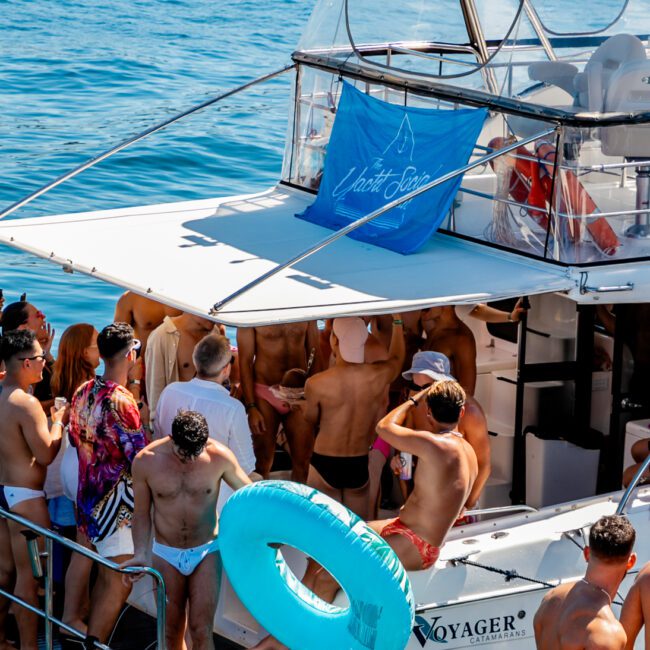 A group of people in swimwear are gathered on the deck of a white yacht named "Voyager" on a sunny day. Some are standing, chatting, while others appear to be preparing to swim. A large blue flotation ring is prominent in the foreground. The sea is calm and boats are visible in the background, perfect for Sydney Harbour Boat Hire.