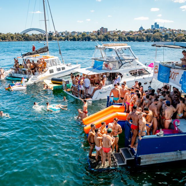 A lively boat party on a sunny day featuring several yachts from The Yacht Social Club Sydney Boat Hire and people swimming in the water. Guests are socializing, dancing, and lounging on inflatable floats. The background includes city buildings and a blue sky with scattered clouds.