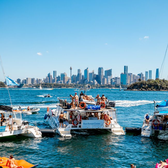 Boats filled with people are anchored in a sunny harbor with a city skyline in the background. Several people are floating and swimming, enjoying the vibrant scene on a clear day. Inflatable pool toys and kayaks add to the fun, creating the perfect atmosphere for Boat Parties Sydney The Yacht Social Club.