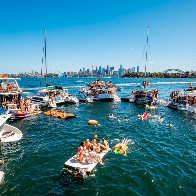 A lively scene of yachts and boats anchored together on clear blue water features many people swimming and lounging on inflatables. Sydney’s skyline and the Sydney Harbour Bridge are visible in the background under a bright, sunny sky, perfect for The Yacht Social Club Event Boat Charters.