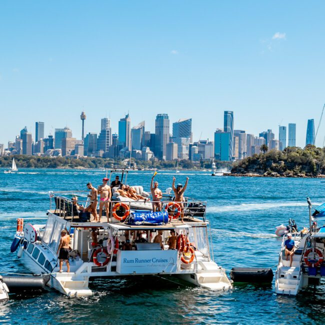 People enjoying a sunny day on a boat named "Rum Runner Cruises" in the middle of a body of water. Some people are swimming near the boat while others lounge on inflatables. The skyline of a modern city with tall buildings is visible in the background, making it perfect for Boat Parties Sydney The Yacht Social Club.