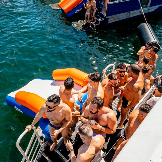 A lively scene at The Yacht Social Club Sydney Boat Hire's party with many shirtless men enjoying the sunny weather. Some people are on the yacht, while others are in the water using colorful inflatables. A staircase leads from the yacht to the water, and other nearby boats are visible.