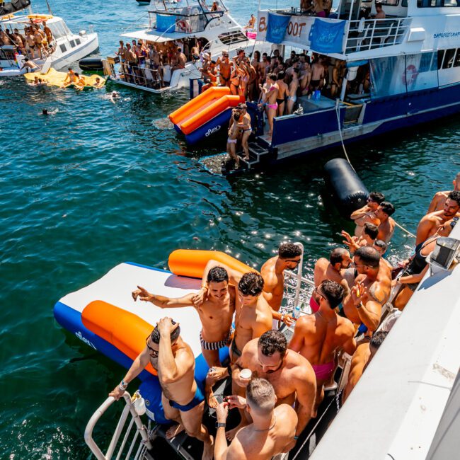 A vibrant scene of a yacht party on clear blue water. Multiple yachts, courtesy of The Yacht Social Club Sydney Boat Hire, are anchored close to each other, packed with people in swimwear enjoying the sunny day. Some are diving into the water, while others are lounging and socializing on the boats.