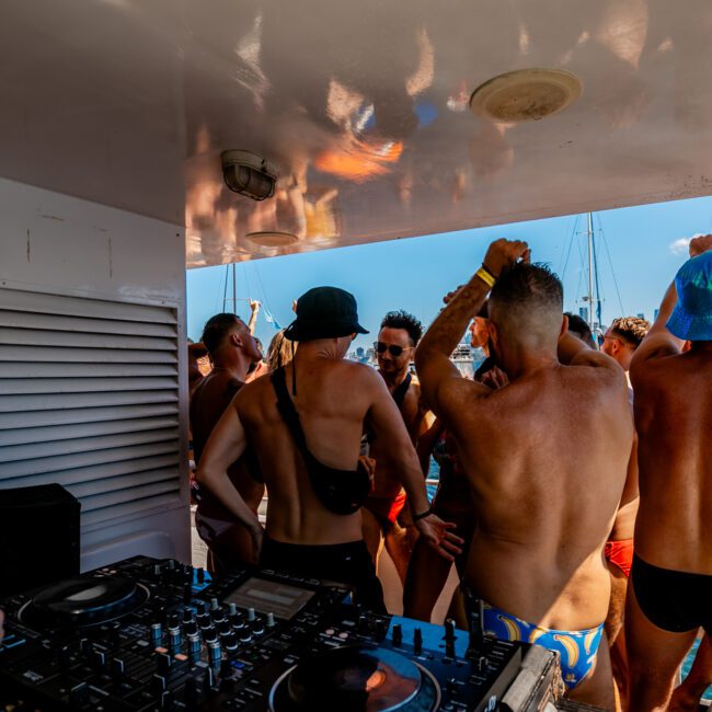 A group of people in swimwear dancing and socializing on a boat under sunny skies, typical of The Yacht Social Club Sydney Boat Hire. A DJ is playing music with a professional setup in the foreground. Several inflatable boats and yachts can be seen on the water in the background.