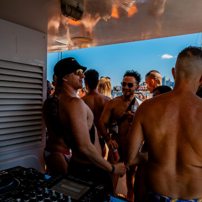 A lively yacht party with shirtless men conversing and holding drinks. The DJ equipment is visible in the foreground, set against the backdrop of a clear blue sky. Everyone seems to be enjoying the sunny day on the water, embracing the luxurious vibe of Sydney Boat Rental and Parties by The Yacht Social Club.