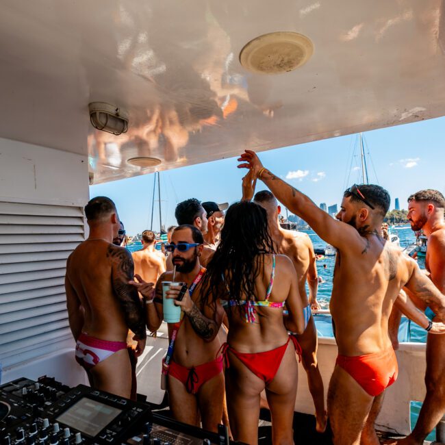 A group of people in swimsuits are dancing and socializing on a luxury yacht with a DJ at the helm. The background shows a sunny day with a clear blue sky and several boats on the water. Some individuals are raising their hands and holding drinks, embodying the spirit of Boat Parties Sydney The Yacht Social Club.